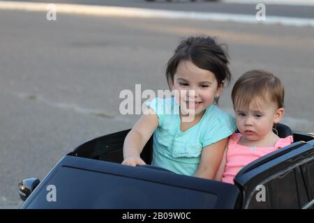 Deux superbes filles vont dans une grande voiture jouet sur l'asphalte de la rue de la ville. Conduite en plein air dans une attraction estivale pour les enfants Banque D'Images