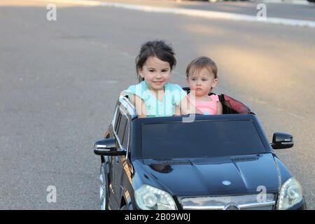 Deux superbes filles vont dans une grande voiture jouet sur l'asphalte de la rue de la ville. Conduite en plein air dans une attraction estivale pour les enfants Banque D'Images