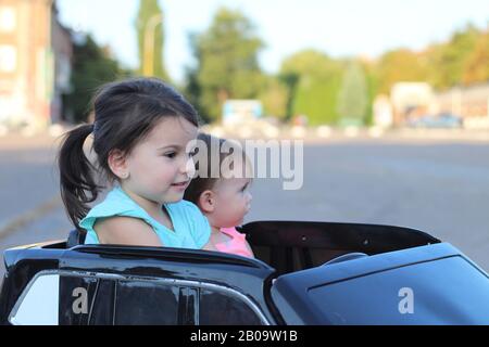 Deux superbes filles vont dans une grande voiture jouet sur l'asphalte de la rue de la ville. Conduite en plein air dans une attraction estivale pour les enfants Banque D'Images