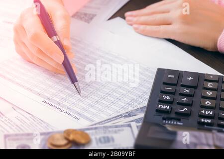 Gros plan des mains de la comptable féminine au bureau qui tient un stylo et recherche des données dans une table. Pile de documents, calculatrice et dollar Banque D'Images