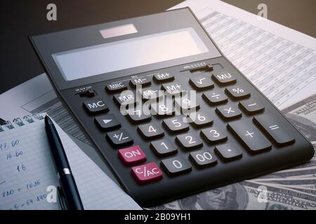 Vue latérale d'une calculatrice comptable noire, d'un rapport avec une table, d'un carnet ouvert, d'un stylo et de plusieurs centaines de dollars de factures sur une table en bois. Bénéfice Banque D'Images