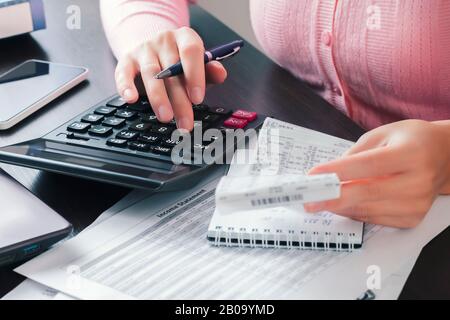 La fille comptable au bureau tient un chèque de caissier dans ses mains et fait des calculs sur une calculatrice, écrivant les résultats dans un bloc-notes. Banque D'Images