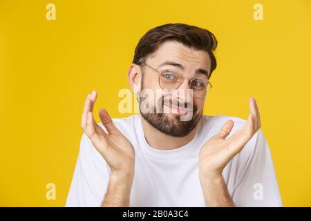 Gros plan de jeune homme regardant et montrant le visage drôle à l'appareil photo, portant le verre. Vue isolée sur fond jaune. Banque D'Images