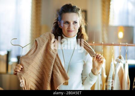 pensive moderne 40 ans femme en chandail blanc et jupe dans la maison moderne en hiver ensoleillé choix des chandails suspendus sur un porte-vêtements en cuivre. Banque D'Images
