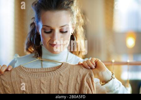 élégante femme de ménage de taille moyenne dans un pull blanc et une jupe dans le salon moderne en hiver ensoleillé tenant des chandails sur un cintre en cuivre. Banque D'Images