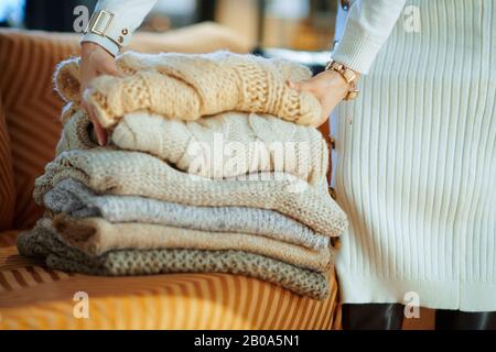 Gros plan sur la femme d'âge moyen en chandail blanc et jupe dans la maison moderne en hiver ensoleillé à proximité des chandails pliants de canapé pour le stockage. Banque D'Images