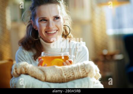 femme souriante d'âge moyen en sweat blanc et jupe avec tas de chandails et tasse de thé avec citron regardant l'espace de copie dans la vie moderne Banque D'Images