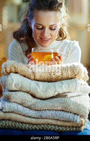 élégante et décontractée, la femme de ménage de 40 ans est en sweat blanc et jupe avec une pile de sweaters à déguster une tasse de thé avec du citron à la maison moderne en hiver ensoleillé Banque D'Images