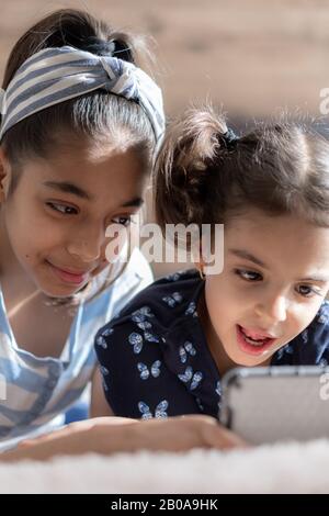 Deux jeunes filles à peau foncée, sœurs, sont couchées au lit et regardent un smartphone sur le fond du soleil depuis la fenêtre. Filles persanes Banque D'Images