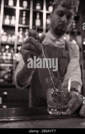 Un animateur de cuisine démodé au bar avec un manche de bain très long, en verre, tête en photo à droite - chemise blanche, tablier, bouteilles sur étagères Banque D'Images