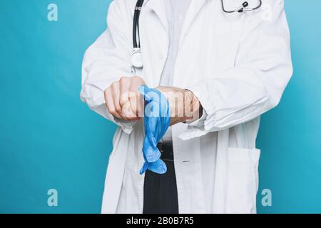 La photo de récolte de l'homme médecin en manteau blanc met sur des gants médicaux en caoutchouc isolés sur le fond bleu Banque D'Images