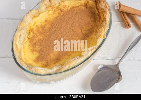 Tarte au lait rustique sur une table blanche avec des bâtons de cannelle et une cuillère de service - photo sélective du dessert sud-africain traditionnel Banque D'Images