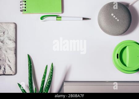 Concept d'assistant numérique. Enceinte plate avec éclairage LED activée avec smartphone court, bloc-notes vert, stylo, usine aloe vera et tasse à café réutilisable. Banque D'Images