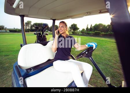 Un golfeur féminin parle au téléphone tout en étant assis dans une voiturette de golf. Banque D'Images