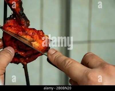 Un chef coupe dans une pièce cuite de poulet mariné tandoori sur une brochette en métal dans une cuisine. Banque D'Images