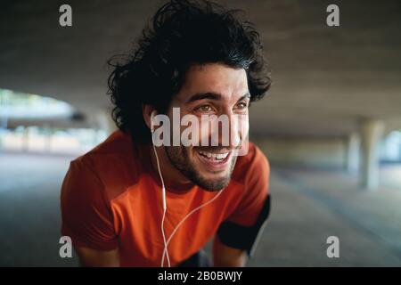 Gros plan portrait d'un jeune homme heureux épuisé avec écouteurs dans ses oreilles prenant la pause après le jogging dans le parc - résolutions de nouvelle année Banque D'Images