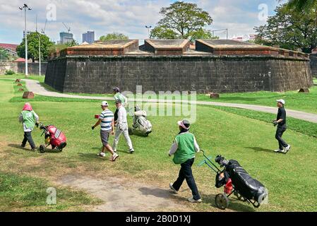 Manille, Philippines: Un groupe de golfeurs asiatiques avec des caddies femelles marchant sur le terrain de golf en face du mur d'Intramuros Banque D'Images