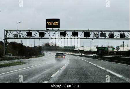 Une voiture conduit le long d'une autoroute britannique, sous de fortes pluies, avec un panneau sur un bras au-dessus de l'autoroute qui conseille aux automobilistes de ralentir pour l'eau de surface. Banque D'Images