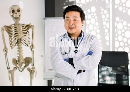 Portrait d'un sympathique médecin coréen asiatique à son bureau souriant à l'appareil photo. Jeune clinicien souriant, blouse qui croise les bras sur la poitrine, humain Banque D'Images
