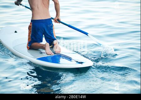 Jeune homme Ayant du plaisir à Pagayer debout dans la mer. SUP. Guy Training le matin sur le panneau de Paddle près des rochers. Banque D'Images