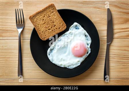 L'oeuf frit se rapproche dans une plaque noire sur une table en bois avec pain grillé et ustensiles. Petit-déjeuner. Côté ensoleillé oeuf doux frit Banque D'Images