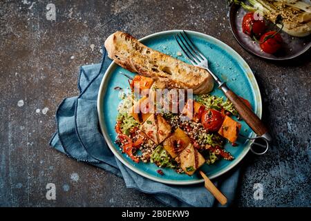 Deux brochettes de légumes grillés servies avec une salade de quinoa verte et une baguette grillée sur une plaque bleue dans une vue de dessus sur un fond gris rustique Banque D'Images