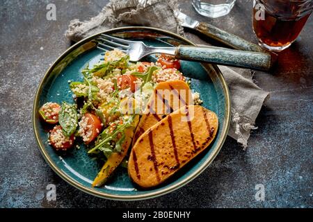 Des tranches de patate douce au barbecue, servies avec de la salade de quinoa et de roquette et des boissons sur un fond rustique pour un délicieux repas végétalien et végétarien Banque D'Images