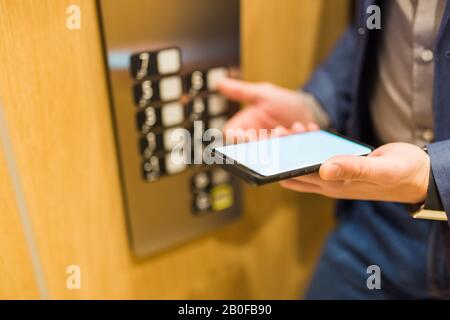 Mains de l'homme tenant un téléphone portable à écran vide lors de l'utilisation du panneau de commande de l'élévateur. Banque D'Images