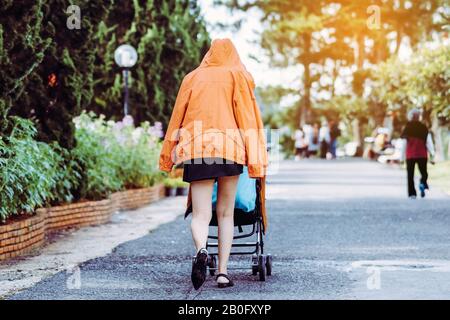 Vue arrière de la femme dans une veste à capuche orange et une jupe courte noire avec des promenades à poussettes pour bébé pour faire de l'exercice dans le jardin. Banque D'Images