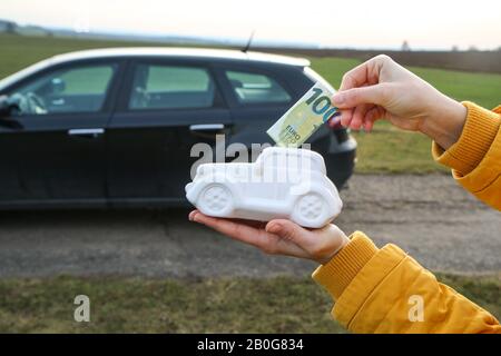 La main entre un billet dans la boîte à monnaie en forme de voiture en céramique. La vraie voiture est derrière. Il peut être le symbole des coûts des réparations, des investissements, Banque D'Images