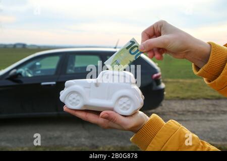 La main entre un billet dans la boîte à monnaie en forme de voiture en céramique. La vraie voiture est derrière. Il peut être le symbole des coûts des réparations, des investissements, Banque D'Images