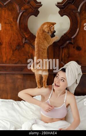 jeune femme avec un chat rouge sur son épaule, serviette sur sa tête repose dans un vieux lit, souriant. Banque D'Images