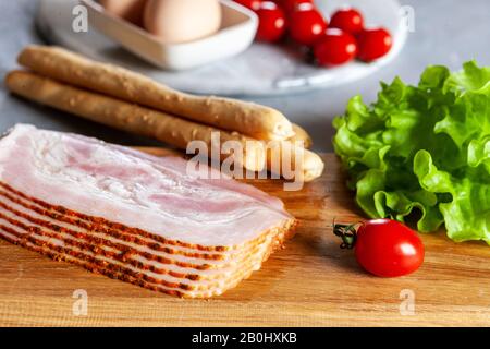 Bacon pancetta traditionnel servi avec des olives marinées, des tomates, des feuilles de salade verte, des œufs et du pain de grissini, des ingrédients pour le petit-déjeuner. Gros plan, haut Banque D'Images