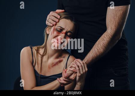 L'homme a mis sa main sur la tête féminine, comme embrasser. Ils tiennent les mains. Victime avec ecchymoses assis à proximité. Fond bleu. Violence familiale. Gros plan. Banque D'Images