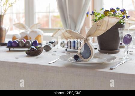 Décoration festive de table de Pâques, serviettes en forme d'oreilles de lapin, œufs de poule et de caille teints, gâteaux, violettes potées, plumes, dîner en famille o Banque D'Images