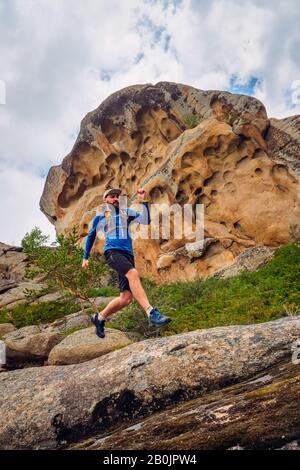 Athlète Guy qui longe un sentier de montagne. Sentier Qui S'Exécute dans les montagnes parmi les rochers. L'homme en jersey bleu et short noir s'entraîner à l'extérieur Banque D'Images