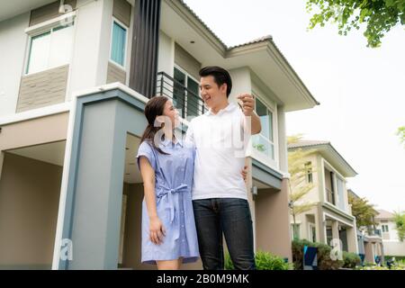 Portrait de jeune couple asiatique debout et embrassant ensemble et tenant la clé de maison regardant heureux devant leur nouvelle maison pour commencer la nouvelle vie. Famille Banque D'Images