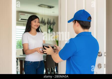 Livraison asiatique jeune homme dans un sourire bleu uniforme et tenant des boîtes de nourriture en face de la maison et la femme asiatique acceptant une livraison de boîtes de nourriture de livym Banque D'Images