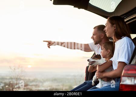 Vue latérale de la famille assise dans le coffre de voiture à l'extérieur de la ville, en regardant le coucher du soleil, le père pointe à l'horizon, copier l'espace Banque D'Images