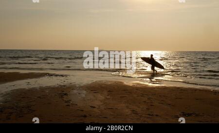 Silhouette d'homme de surf avec planche de surf sur la surface de l'eau. Surf sur la plage au coucher du soleil. Sports nautiques en plein air aventure style de vie.activité d'été. Banque D'Images