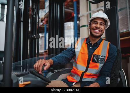 Jeune conducteur de chariot élévateur assis dans le véhicule en entrepôt souriant regardant la caméra Banque D'Images