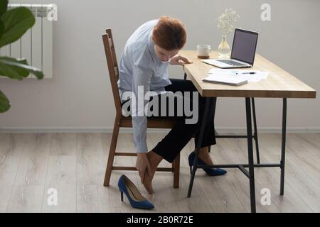 Femme d'affaires fatiguée assise sur une chaise de bureau et massant le pied, se sentant inconfortable après avoir porté des chaussures avec des talons, des muscles tendus, employé se faisant facilement e Banque D'Images