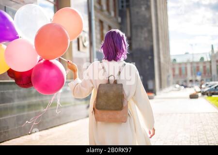 Fille aux cheveux violets, dans un gilet blanc et un sac à dos beige, avec un tas de ballons de couleur, debout la tourner en arrière devant le fond gris mur o Banque D'Images