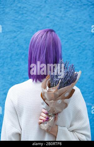 Jeune fille avec des cheveux violets, chemisier blanc, debout, tourner son dos avec le bouquet de lavande dans le papier enveloppé dans sa main droite devant le dos de mur bleu Banque D'Images