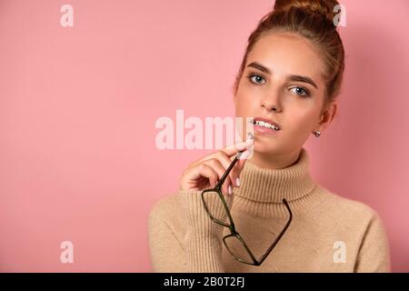 Une fille avec une peau propre et un grand pain se tient sur le fond rose et regarde soigneusement dans le cadre, tenant les verres par l'oreille. Banque D'Images