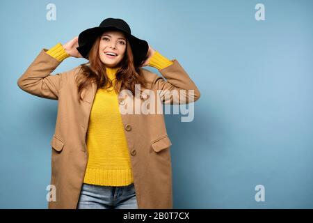 Une belle brunette dans un manteau et un chapeau noir se tient sur un fond bleu avec ses mains derrière sa tête et sourire. Banque D'Images