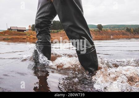 Jambes en pantalon et bottes imperméables marchant mi-courant dans une rivière peu profonde. Banque D'Images