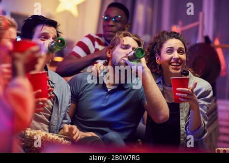 Les jeunes hommes boivent de la bière à partir de bouteilles tout en étant assis sur un canapé avec des filles et en riant pendant la fête Banque D'Images