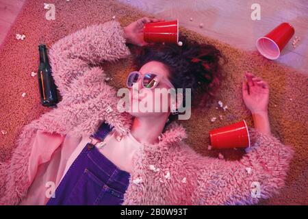 Jeune femme dans des lunettes de soleil dormant sur le sol entre des verres vides et des bouteilles après la fête Banque D'Images