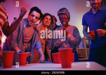 La femme africaine souriante est très excitée tout en jouant dans le jeu de bière-pong avec ses amis pendant la fête Banque D'Images